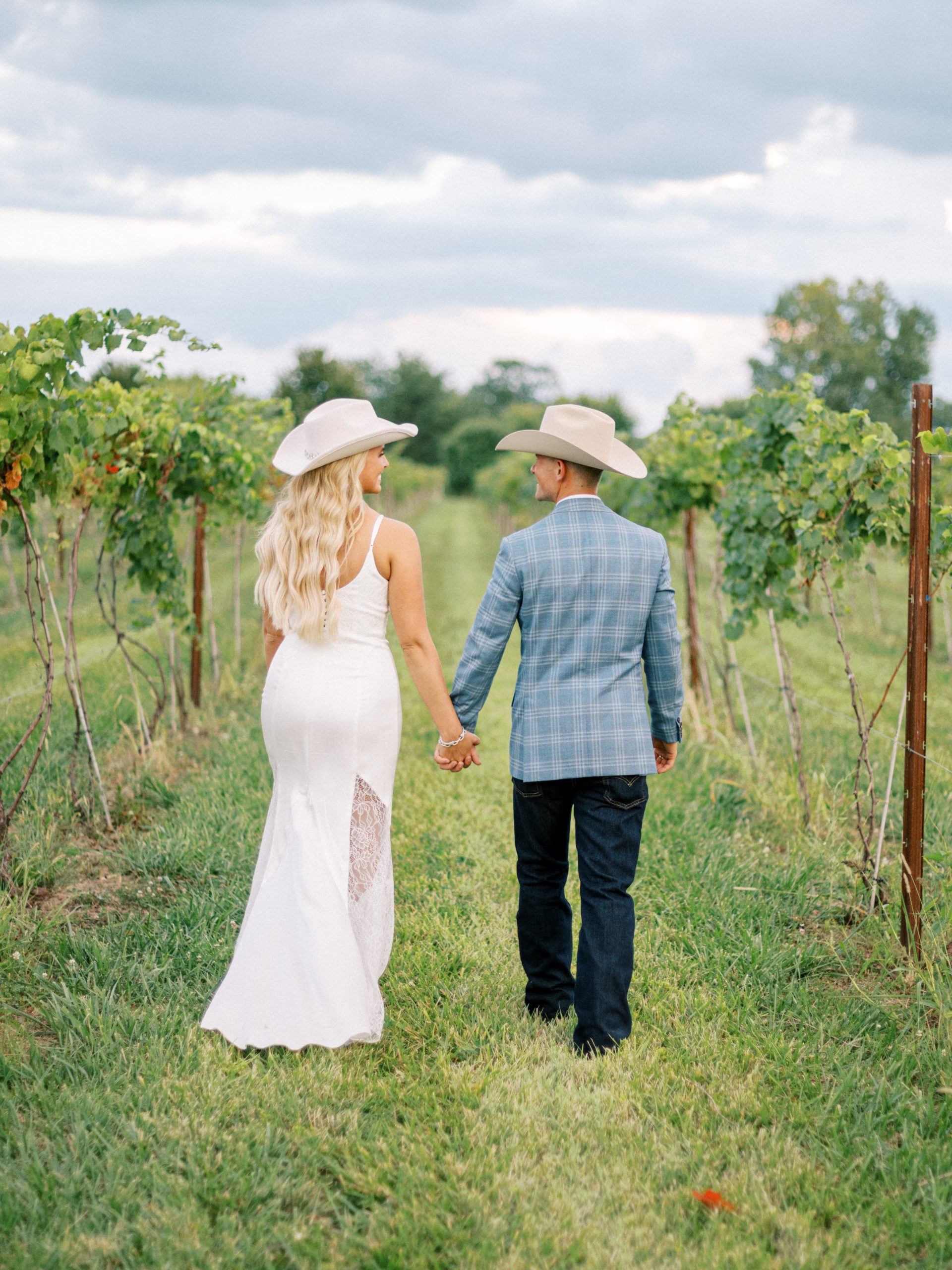 Bride and groom wearing cowboy hats walking hand in hand through vineyard, backs to camera at Retreat 21 near Columbus, Ohio